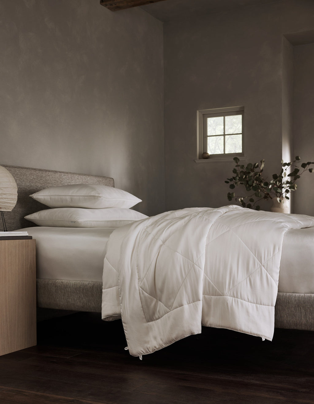 A neatly made bed features the Cozy Earth Bamboo Viscose Comforter with white bedding in a modern, softly lit bedroom. A nightstand with a lamp and plant sits beside the bed, while a small window allows natural light in.|Filling:Standard 